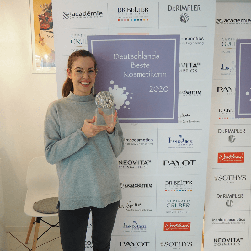 Louisa Galland gewinnt den Preis ‚Deutschlands beste Kosmetikerin 2020‘ und hält stolz ihre Auszeichnung vor der offiziellen Sponsorenwand.